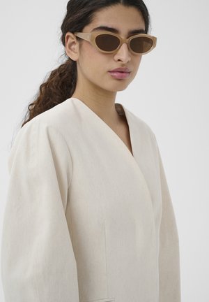 Žena s tmavými kučeravými vlasmi v nízkom copu, oblečená v béžovom oversized saku a svetlohnedých slnečných okuliaroch typu cat-eye, neutrálne pozadie.