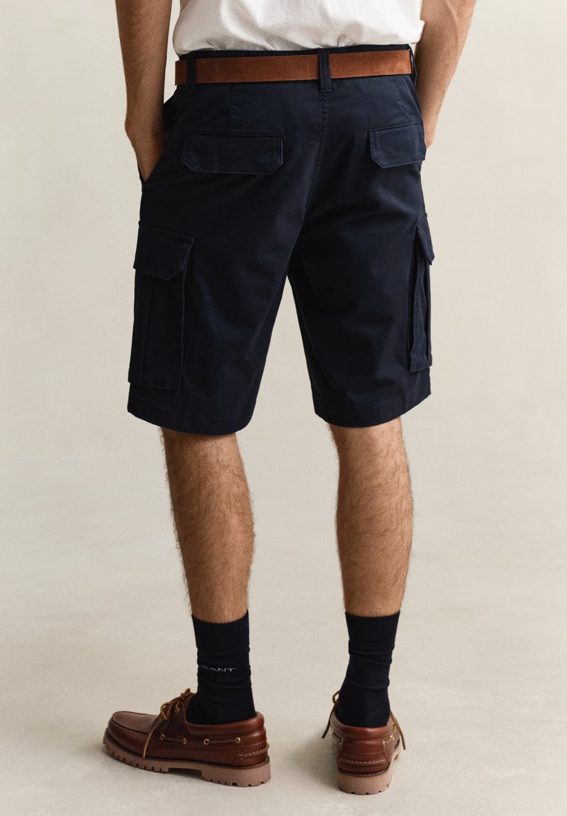 Uomo che indossa pantaloncini cargo blu navy con molte tasche, cintura marrone, calzini neri e scarpe stringate marroni, in piedi contro uno sfondo neutro.