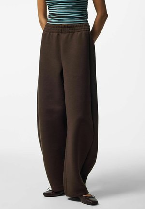 Personne portant un pantalon large marron foncé ample, des ballerines noires et un haut sans manches rayé vert et noir, debout sur un fond blanc.