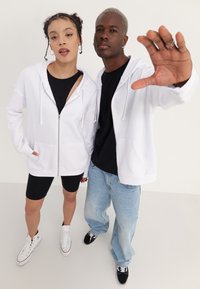 Due persone stanno insieme, entrambe con felpe bianche con zip. Lo sfondo è semplice. La donna ha la pelle chiara e indossa un top nero, mentre l'uomo ha la pelle scura e indossa una maglietta nera con dei jeans.