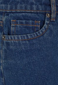 Gros plan d'une poche de jean en denim bleu avec des coutures orange et des rivets métalliques.
