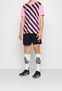 Maglia da calcio a righe nere e rosa con scollatura a V, abbinata a pantaloni corti neri e calze bianche con motivi geometrici neri, indossata con scarpe da calcio nere.