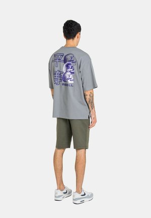 Man wearing gray oversized T-shirt with three purple skulls and Asian characters on back, olive shorts, white sneakers, standing with back to camera.