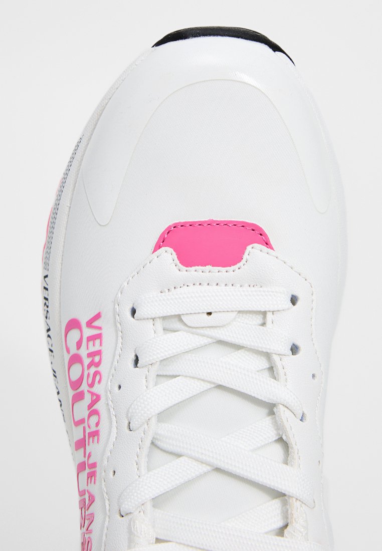 Zapatilla blanca con acentos rosas, diseño con cordones y material texturizado. Presenta el logo "VERSACE JEANS COUTURE" en el lateral.
