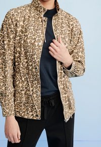 Leopard-print jacket in beige and brown fabric, with a high collar, zip closure, and elastic cuffs; worn over a black top and pants.