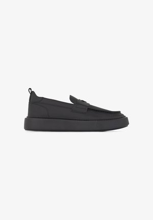 Scarpe slip-on nere con suola in gomma texturizzata, caratterizzate da un design piatto e una cinturina decorativa sulla parte superiore. Materiale in pelle liscia.