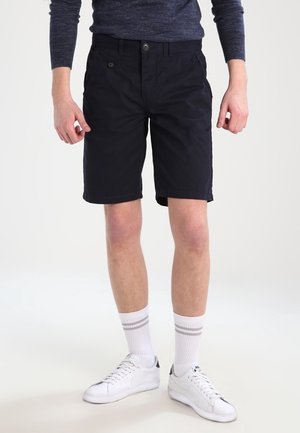 Personne portant un short noir jusqu'aux genoux, des chaussettes blanches à rayures grises, des baskets blanches et un chemisier sombre à manches longues.