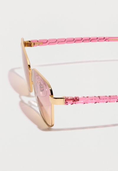 Γυαλιά ηλίου με χρυσή κορνίζα, ροζ χρωματισμένους ορθογώνιους φακούς και διαφανή ροζ βραχίονες με υφή, ακουμπισμένα σε λευκή επιφάνεια.