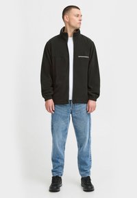 Svart fleecejakke med høy krage og frontlomme med glidelås, kombinert med lys blå denimjeans og svarte joggesko. Enkel, uformell design.