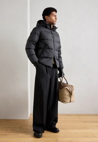 Veste matelassée noire avec capuche, design matelassé et zip à l'avant. Associée à un pantalon large noir et un sac en cuir marron. Gants noirs portés.