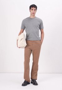 Grijze katoenen T-shirt, tan corduroy broek, zwarte schoenen. Model houdt een crèmekleurige tote bag vast en staat tegen een witte achtergrond.