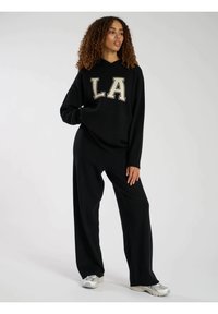 Svart huvtröja med "LA" i beige bokstäver, kombinerad med vida svarta byxor. Modellen står mot en enfärgad bakgrund.