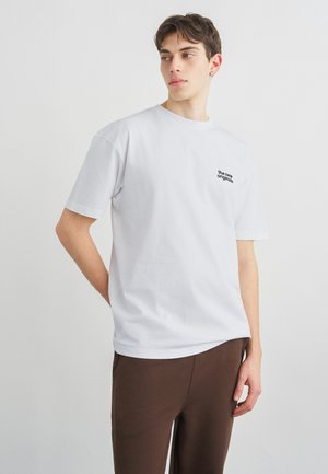 Junger Mann trägt ein weißes T-Shirt mit dem Schriftzug „the new originals“, braune Hose, blickt seitlich vor einem schlichten hellen Hintergrund.