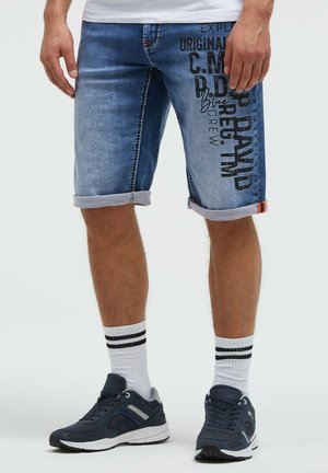 Mann trägt blaue Jeansshorts mit schwarzem Text am rechten Bein, weiße Socken mit schwarzen Streifen und dunkelblaue Sportschuhe.