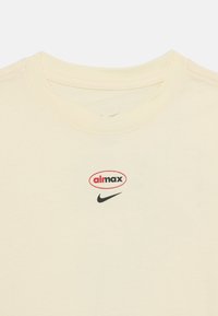 T-shirt en coton beige clair avec un col rond. Présente un logo "aimax" rouge et noir ainsi qu'un swoosh noir de Nike sur le devant. Texture lisse.