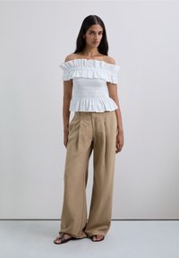 Kvinna står klädd i vit off-shoulder blus med volanger, lösa beige byxor och svarta sandaler mot en enkel ljusgrå bakgrund.