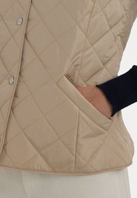 B éžová prošívaná vesta s diamantovým vzorem, boční kapsou a stříbrnými patentními knoflíky. Oděv má hladkou strukturu a zaoblený lem.