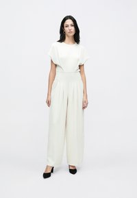 Salopetă crem cu mâneci dolman scurte, corsaj mulat și pantaloni evazați. Material texturat și detalii plisate care îmbunătățesc designul.