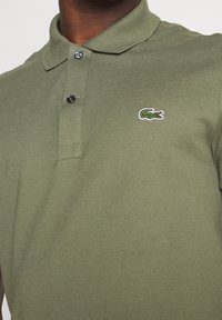 Polo vert olive en tissu texturé, avec une patte de boutonnage à deux boutons et un petit logo crocodile brodé sur la poitrine gauche.