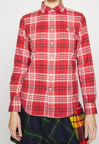 Röd rutig skjorta med vita och svarta detaljer, med krage som knäpps, långa ärmar och en diskret broderad logotyp på bröstet.