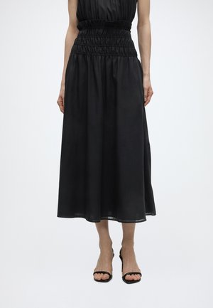 Kobieta ubrana w czarną sukienkę bez rękawów z wcięciem w talii oraz czarne sandały na wysokim obcasie z odkrytymi palcami, stojąca na jednolitym tle.