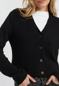 Cardigan nero lavorato a maglia con scollatura a V, polsini e bordo a coste, con sei bottoni sul davanti e un tessuto testurizzato.
