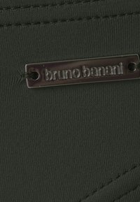 Tamnozelena tkanina s teksturiranom površinom, sadrži srebrnu oznaku ugraviranu s "bruno banani," pričvršćenu uzicom.