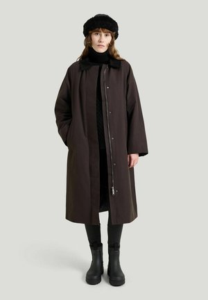 Kvinde iført en lang mørkebrunt frakke, sort sweater, sorte bukser, sorte støvler og en sort pels hat, der står mod en ensfarvet baggrund.