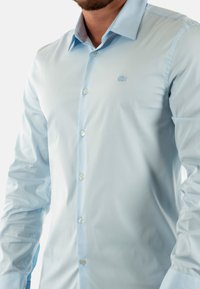 Camisa de manga larga azul claro hecha de una tela suave, con un cierre de botones en la parte delantera y un logotipo sutil en el pecho. Diseño de cuello clásico.