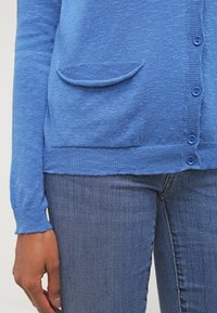 Cárdigan azul claro con textura suave, un bolsillo y cierre de botones, combinado con jeans azul oscuro.