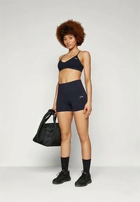 Fiatal nő fekete sportmelltartóban és rövidnadrágban, fekete edzőtáskával a kezében, fekete zoknit és sportcipőt viselve, sima háttér előtt áll.