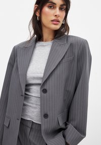 Grå stribet blazer med skræddersyet pasform, der har to knapper, notch-lapper og en enkelt brystlomme. Laget over en lysegrå top.
