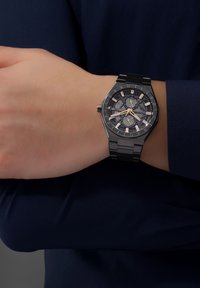 Svart oktagonal klocka med en mörk urtavla och lysande guldiga timmarkörer, en armband av rostfritt stål och tre underklockor för tidtagning.