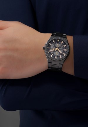 Sort oktagonal ur har en mørk urskive med lysende guld timeindekser, et armbånd i rustfrit stål og tre subskiver til tidsfunktioner.