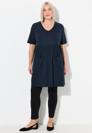 Abito navy a maniche corte con scollatura a V, vita arricciata e due tasche anteriori; abbinato a leggings neri e scarpe flat. Tessuto dalla texture liscia.