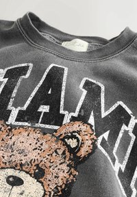 Sweat-shirt gris foncé avec un grand texte "PLAY" blanc et noir et un graphisme texturé d'ours en peluche marron sous le texte, au niveau de la poitrine.