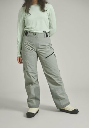 Graue Schneehosen mit lässiger Passform, die mehrere Taschen, schwarze Akzente und kontrastierende Materialpaneele aufweisen. Kombiniert mit weißen Schuhen.