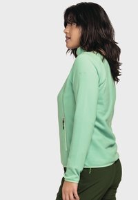 Hellgrüne Fleecejacke mit hohem Kragen, seitlichem Reißverschluss und tailliertem Design. Das Material wirkt weich und hat eine glatte Textur.