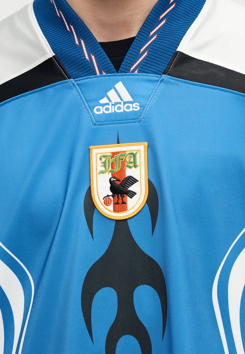 Maillot de sport bleu avec des accents blancs et noirs, logo Adidas au-dessus d'un emblème en forme de bouclier présentant un oiseau noir et les lettres IFA.