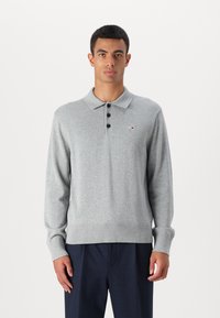 Šviesiai pilkas ilgarankovis polo megztinis, turintis tris mygtukus, įsiūtas rankogalius ir apatinę dalį, su mažu logotipu kairėje krūtinės pusėje.