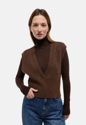 Pull sans manches marron en tricot avec un col en V profond et une texture côtelée, superposé à un col roulé marron ajusté, assorti à un jean bleu taille haute.