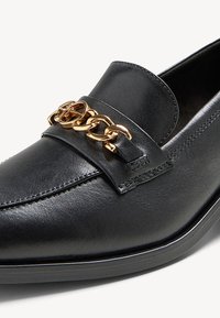 Mocassin en cuir noir avec un accent de chaîne en or sur le cou-de-pied, présentant un design élégant, une texture lisse et un bout arrondi.