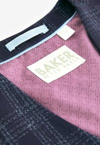 Gros plan d'une doublure de veste violette à motifs avec une étiquette de vêtement blanche "Baker by Ted Baker" et un tissu bleu foncé avec des coutures claires.