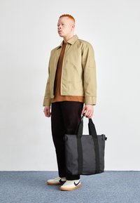 Borsa a tracolla nera con doppi manici, realizzata in materiale liscio e resistente. Il modello indossa un giubbotto marrone chiaro sopra una camicia marrone scuro e pantaloni in velluto a coste.