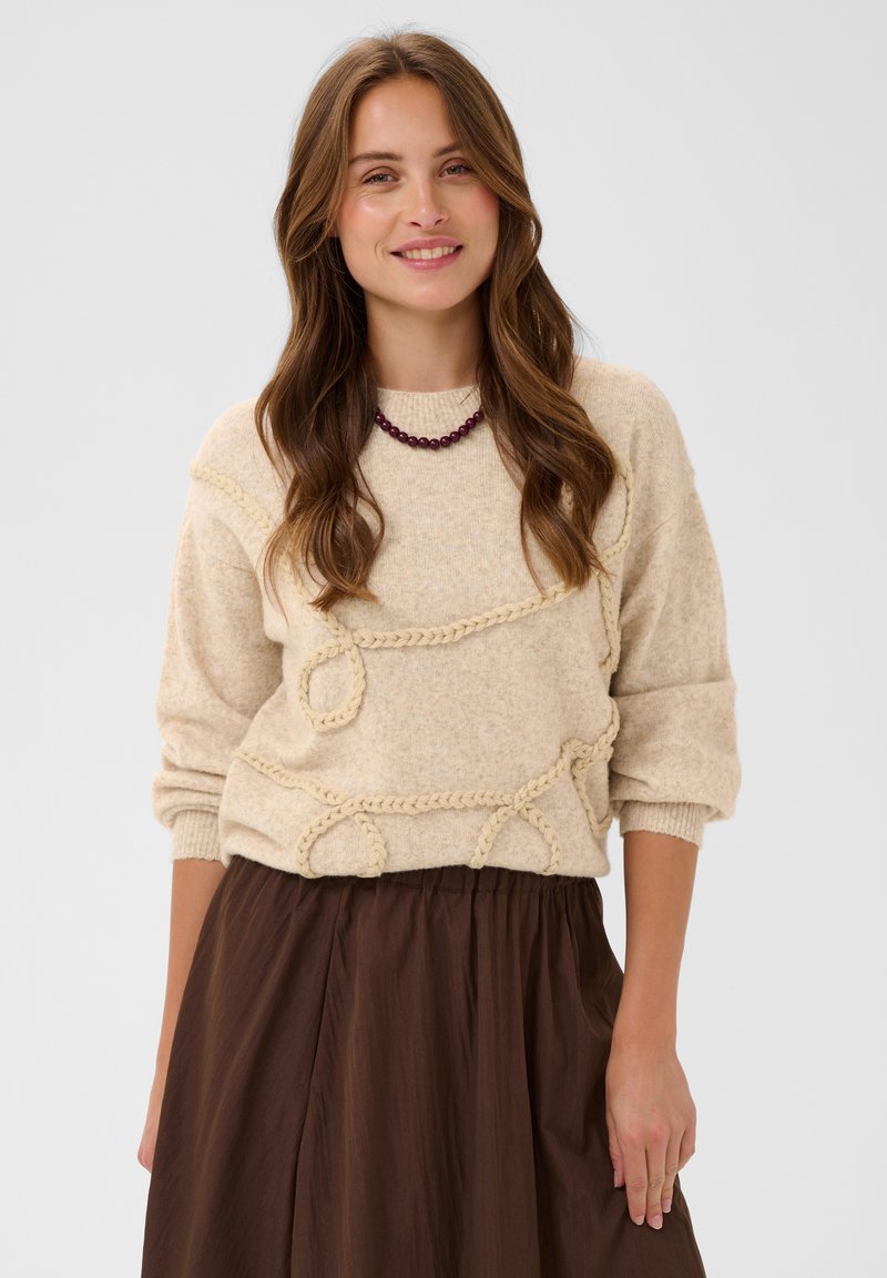 Femme aux longs cheveux bruns porte un pull beige texturé, un collier de perles bordeaux et une jupe marron, souriant devant un fond uni.