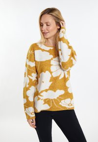 Maglione giallo senape con motivo floreale bianco, colletto rotondo, maniche lunghe e una texture morbida. Sotto sono indossati pantaloni neri.