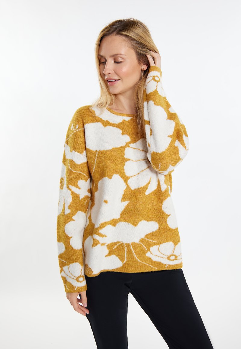 Maglione giallo senape con motivo floreale bianco, colletto rotondo, maniche lunghe e una texture morbida. Sotto sono indossati pantaloni neri.