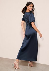 Marineblaues Maxikleid aus Satin mit Puffärmeln, tailliert und mit fließendem Rock. Kombiniert mit silbernen High-Heel-Sandalen und auffälligen Ohrringen.