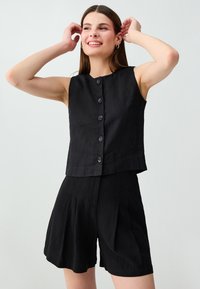 Jeune femme souriante, les mains près de la tête, portant un haut noir sans manches boutonné et un short noir assorti.