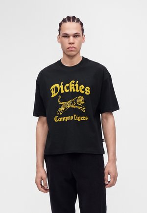 Jeune homme aux cheveux tressés portant un t-shirt noir avec un texte jaune "Dickies Campus Tigers" et un graphisme de tigre sautant.